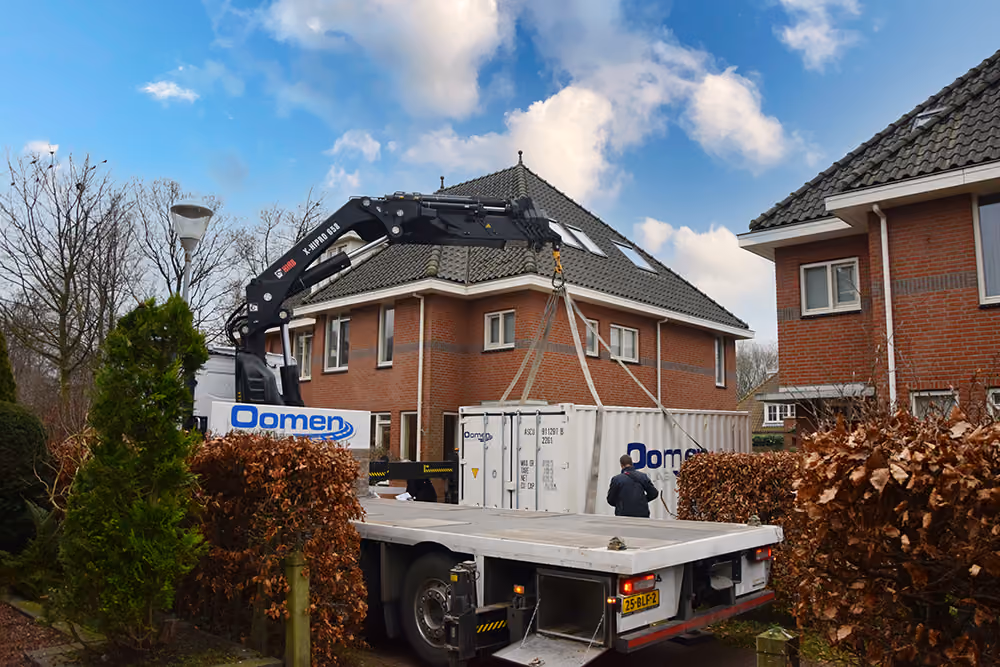 Vrachtwagen van Oomen plaatst met de kraan een opslagcontainer bij een woning